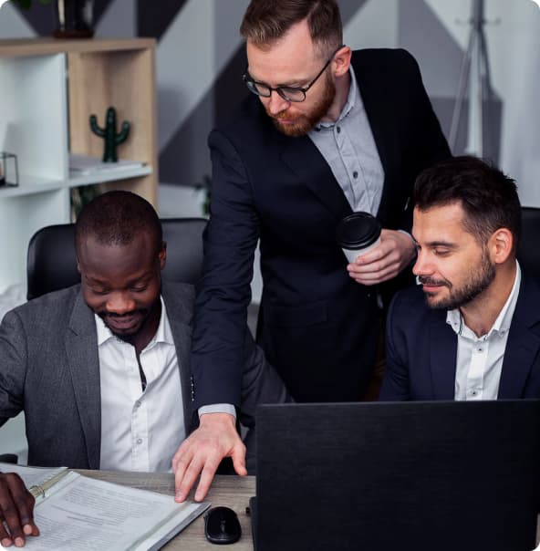 Dois homens sentados, um branco e outro negro, e outro homem em pé observam as informações em um papel sobre uma mesa de escritório com computador e mouse.